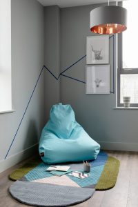 Cork's Marsh's Yard. An image of a corner of a room. In the foreground is a wooden floor with a carpet. In the centre is a aqua coloured bean bag. In the background are the grey walls with 2 framed sketches of cows